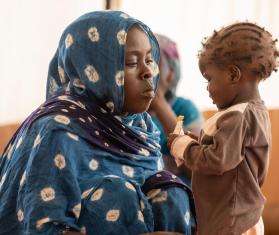 Mother in a blue hijab watches her child eat therapeutic peanut paste in Adré, Chad.