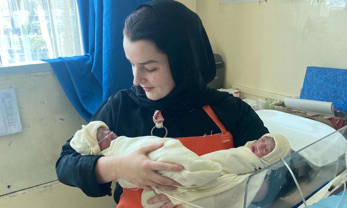 A woman in a head-covering and orange-ish uniform holds a small newborn wrapped in a blanket. 