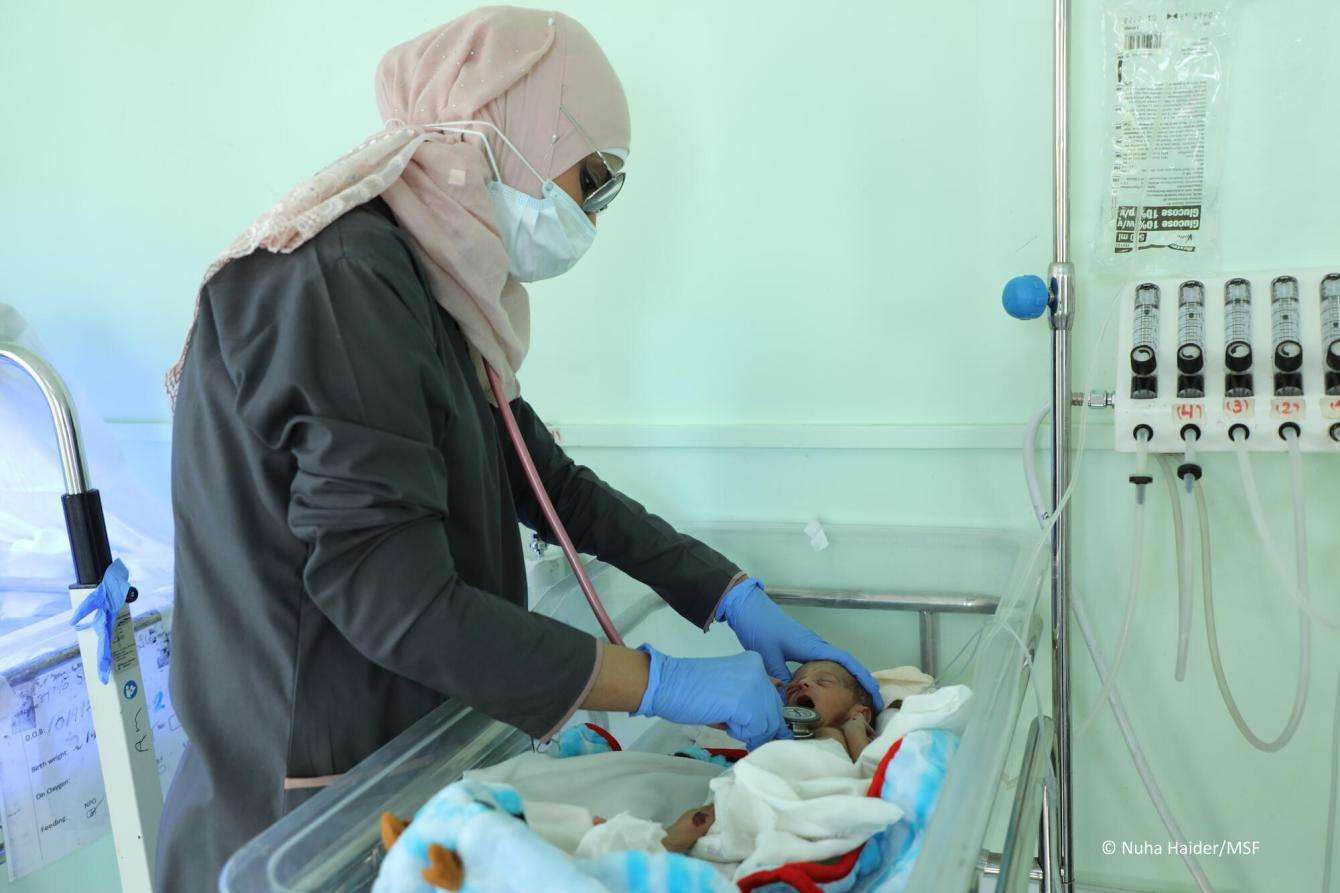 A female medic wearing a mask is using her stethoscope to check a baby who is lying in a medical crib.