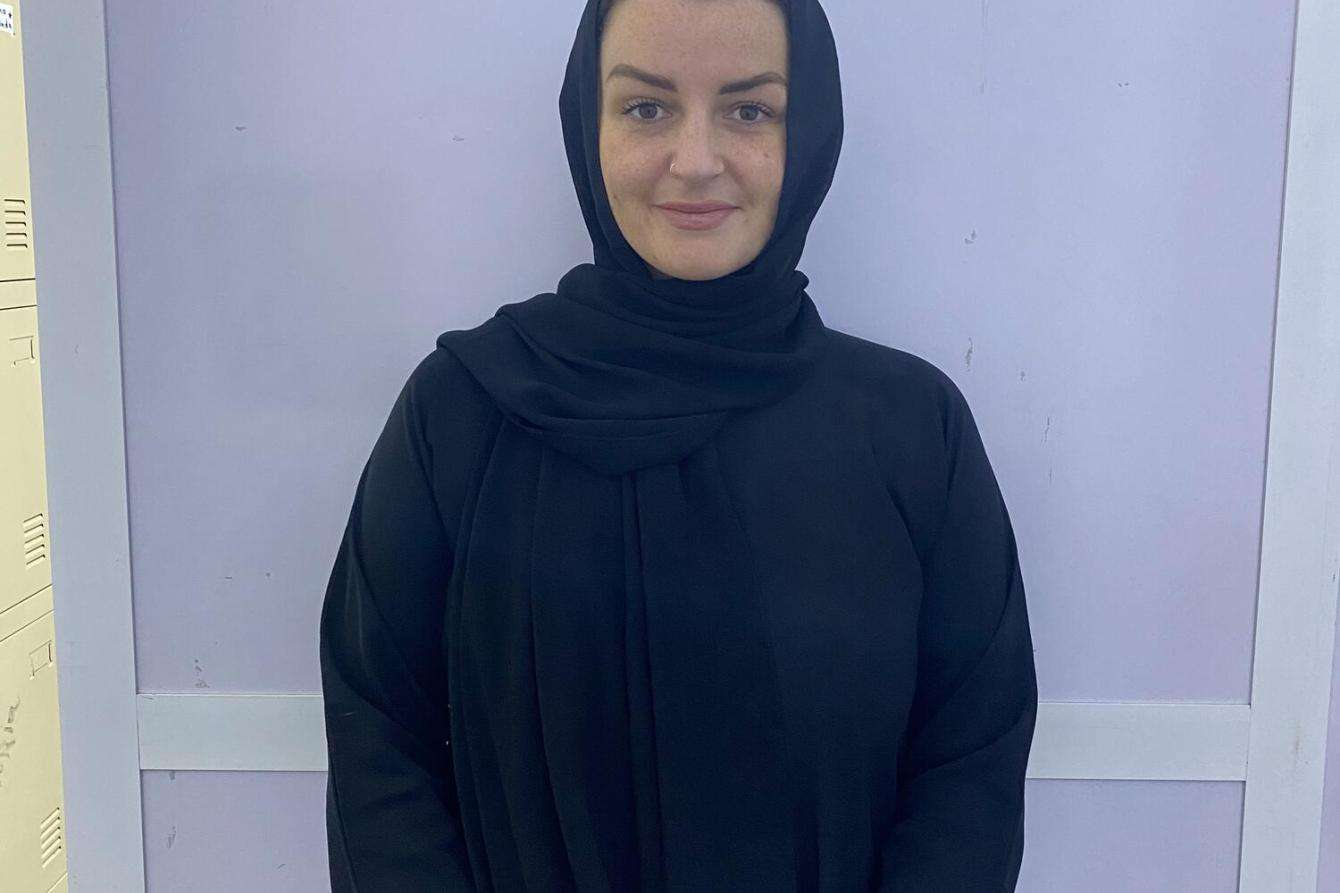 A portrait of a woman with light skin wearing all black, in a hijab, in front of a lavender background.