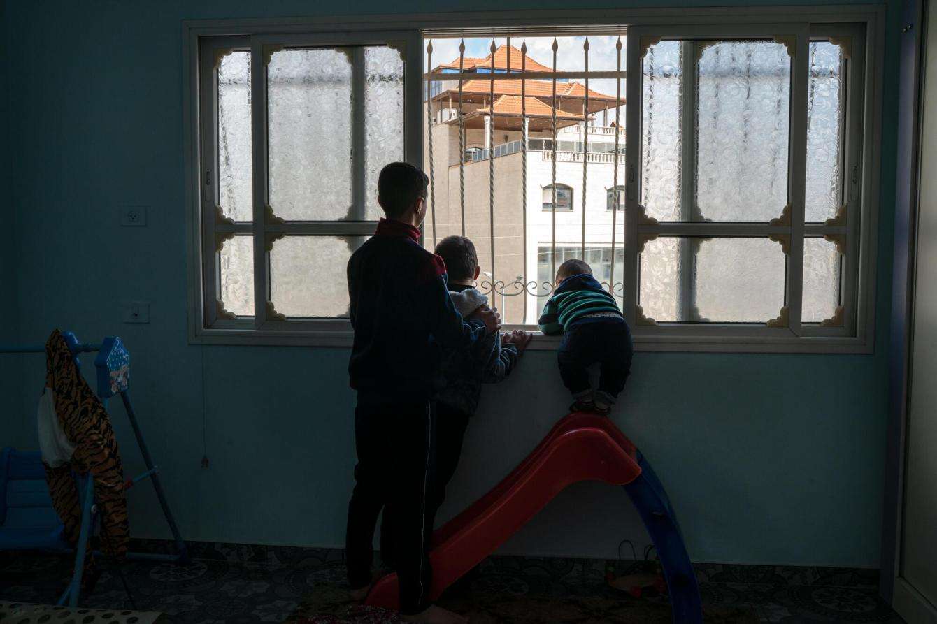 Three Palestinian children stand in a dark room looking through a window in Huwara, West Bank, Occupied Palestinian Territories.