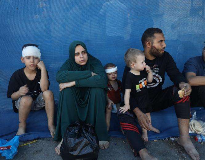 Wounded and displaced Palestinians in Gaza sit in front of a blue wall 