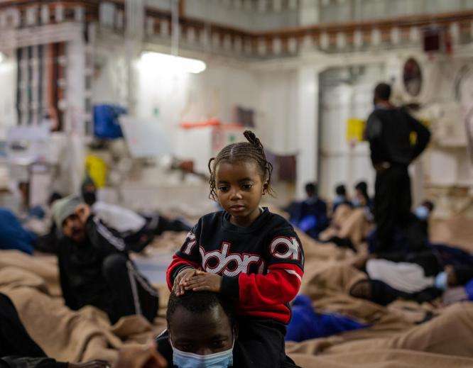 A child stands next to a caregiver on the deck of a search and rescue ship. 