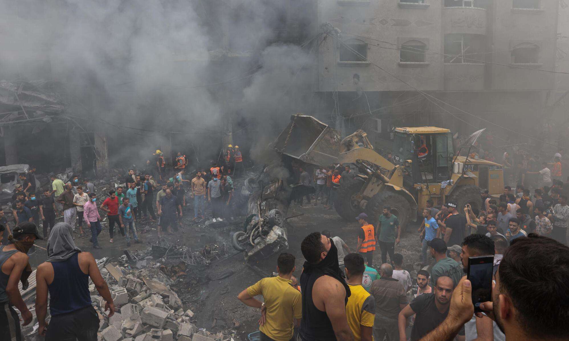A crowd of Palestinians surrounded by rubble and smoke from Israeli airstrikes in Gaza.