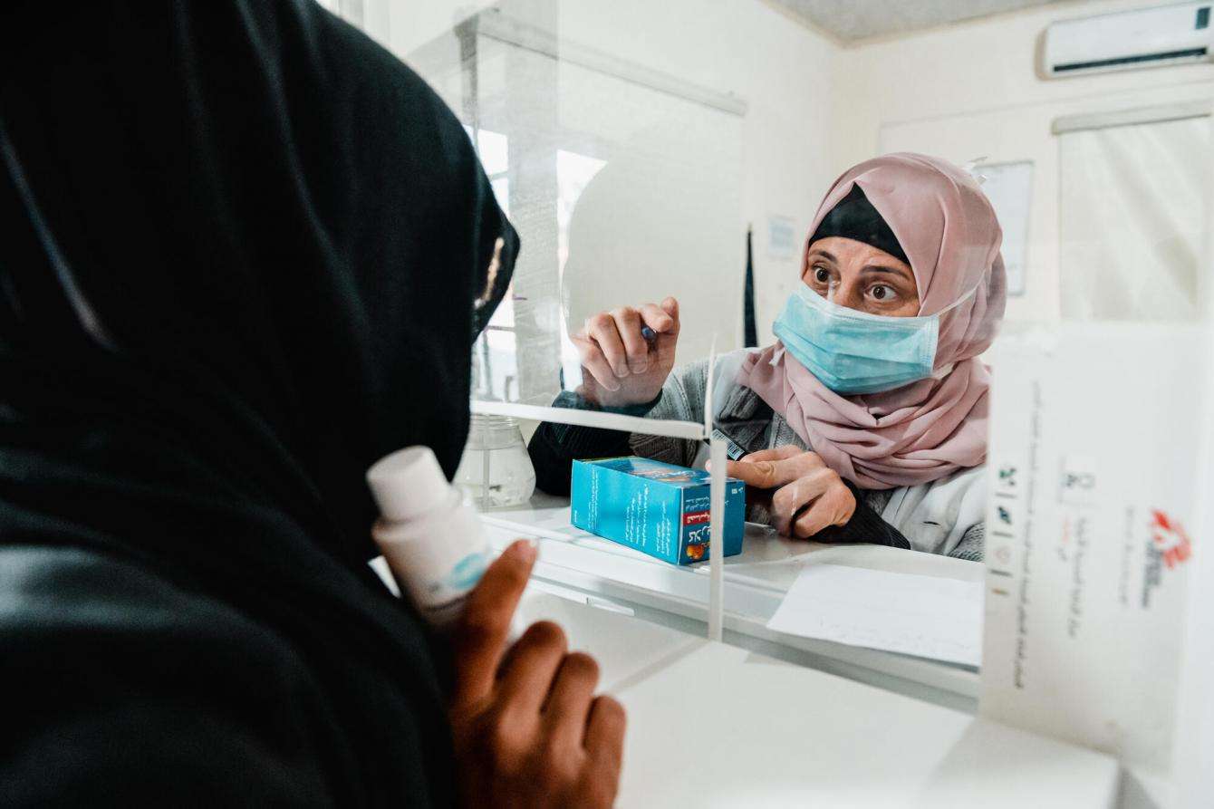 A masked female medic distributes medication to another woman on the other side of the glass.