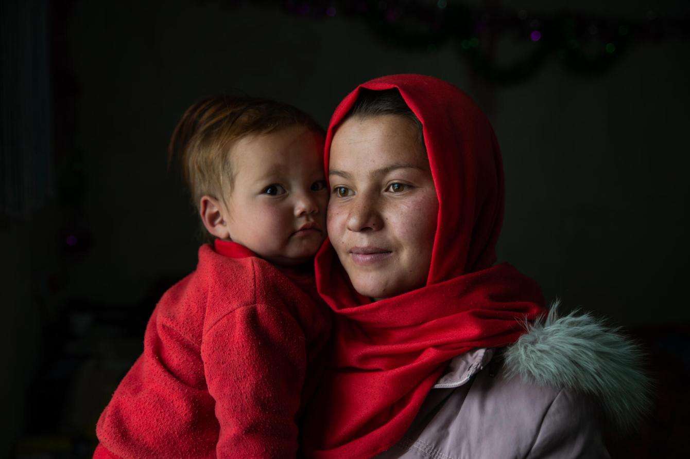 An Afghan young woman in a red headscarf holds her child in a red shirt against a dark background.