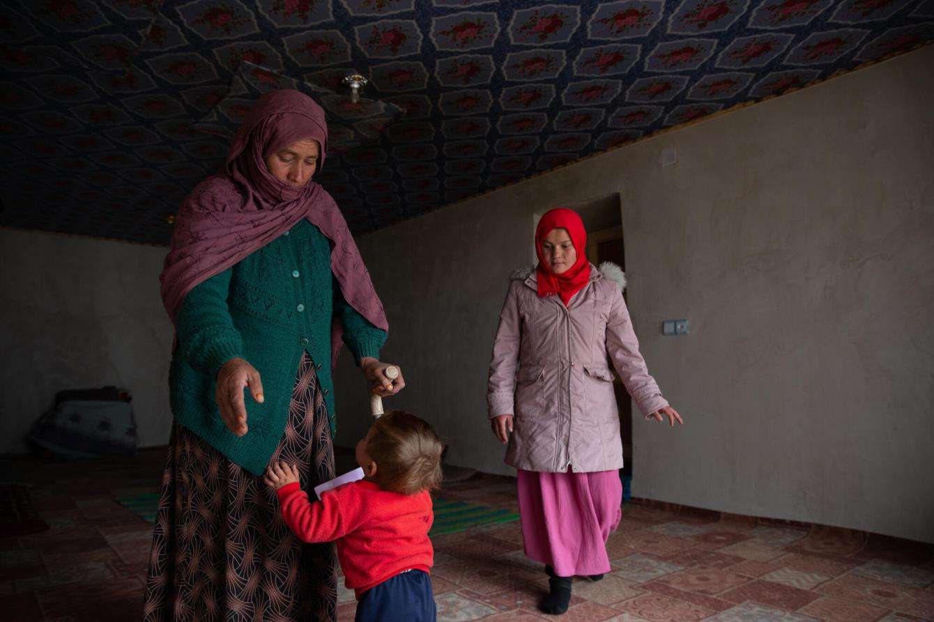 Two women in headscarves play with a child in a red shirt in a dark room in Afghanistan.