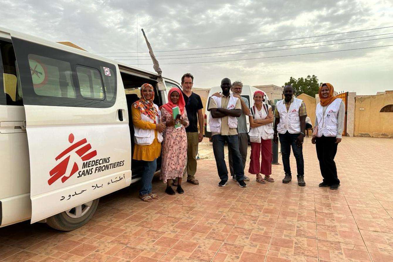 MSF team stands outside vehicle with MSF logo under cloudy sky at Umdawanban hospital in Khartoum state, Sudan.
