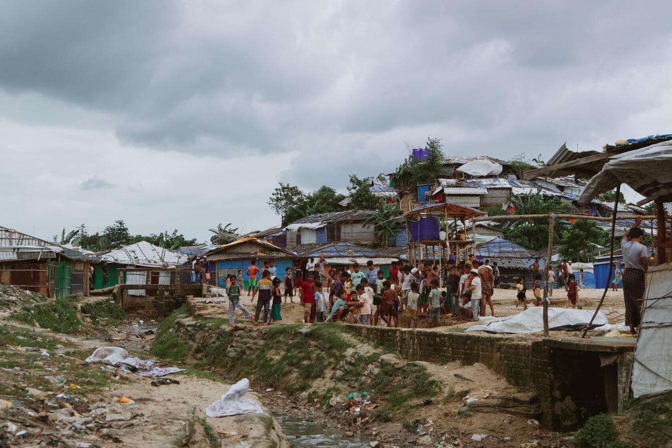Wide-shot of ramshackle houses, some without rooftops, and garbage in the background with dozens of children gathered together in an open space by a stream of water.