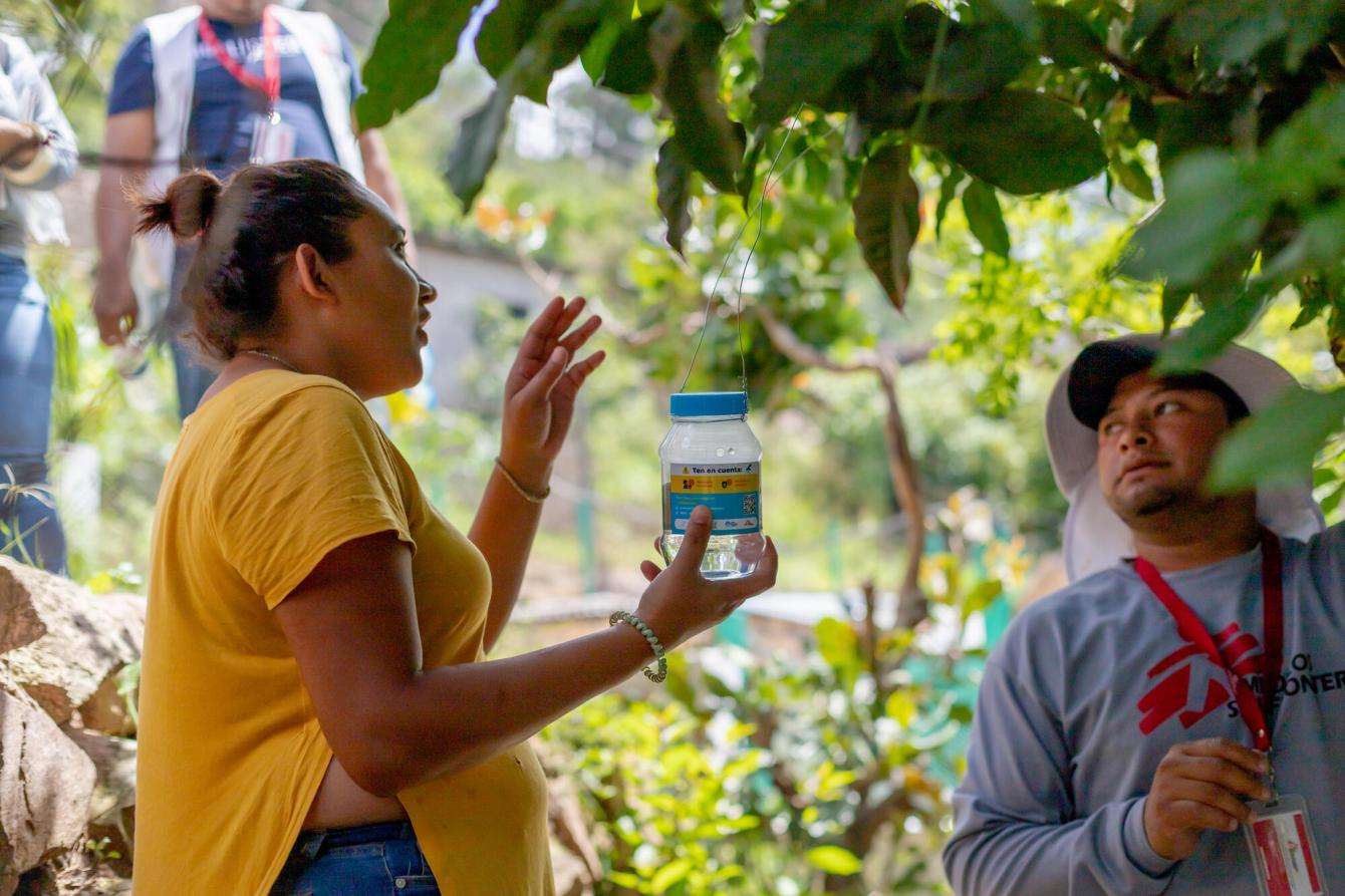 A woman is holding a jar of mosquitos outside, surrounded by trees, speaking to someone who works for MSF.