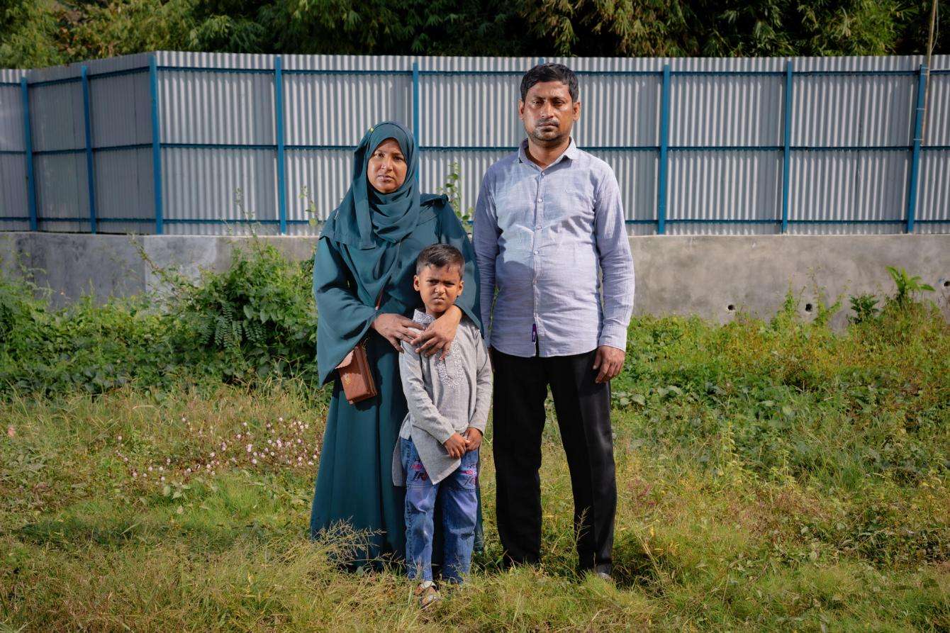 In a grassy area, a woman in a teal hijab has her arms around her young son and her husband in a blue button-up shirt and black plants stands next to them.
