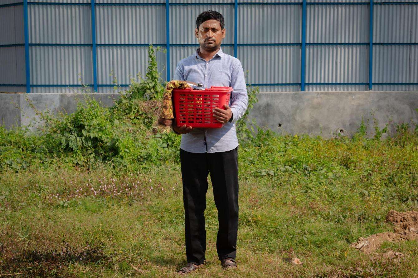 A man stands on the grass holding a red plastic basket full of objects.