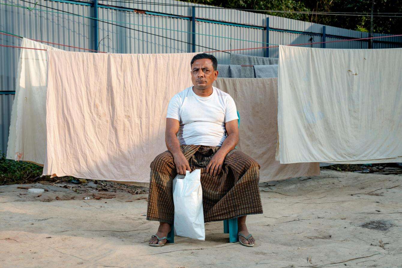 A man in a white T-shirt sits infant of a line of drying white bed sheet with a plastic bag at his feet.