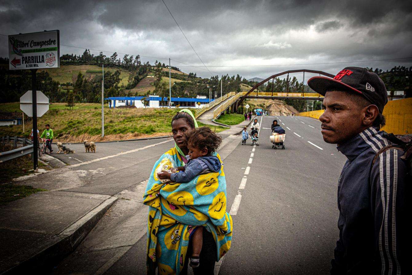 A woman carrying a child and a man travel on a road through the Americas