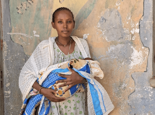 A woman wearing beige dress and scarf holds baby in wrapped in blue fabric against a wall with chipped paint in Tigray, Ethiopia.