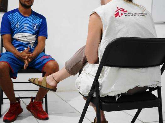 Two people whose faces we cannot see sit on black plastic chairs, facing each other. One is wearing a blue T-shirt the other is in an MSF vest.